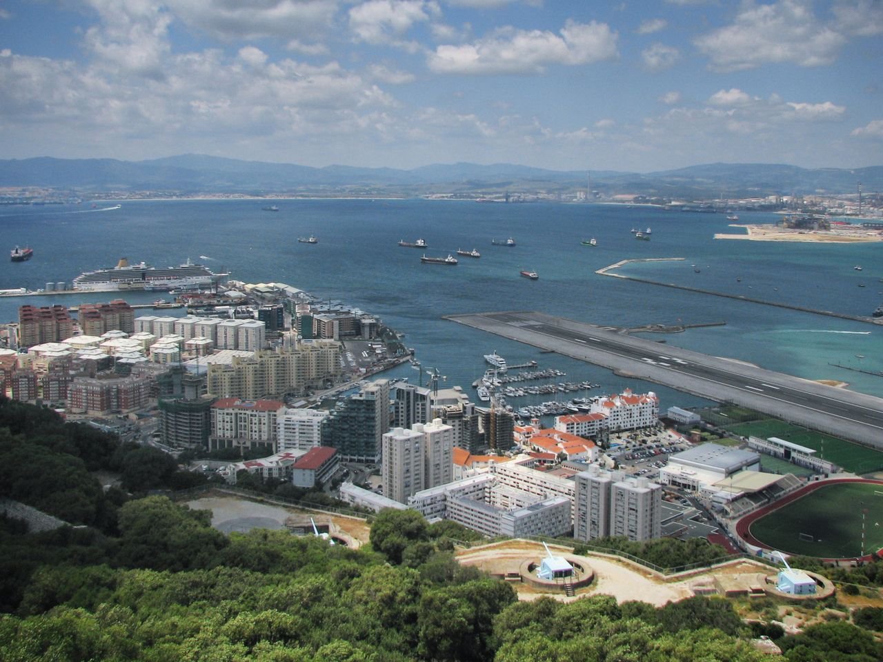Scenic aerial view of a bustling coastal city with a marina, surrounded by mountains and a bright blue sea.